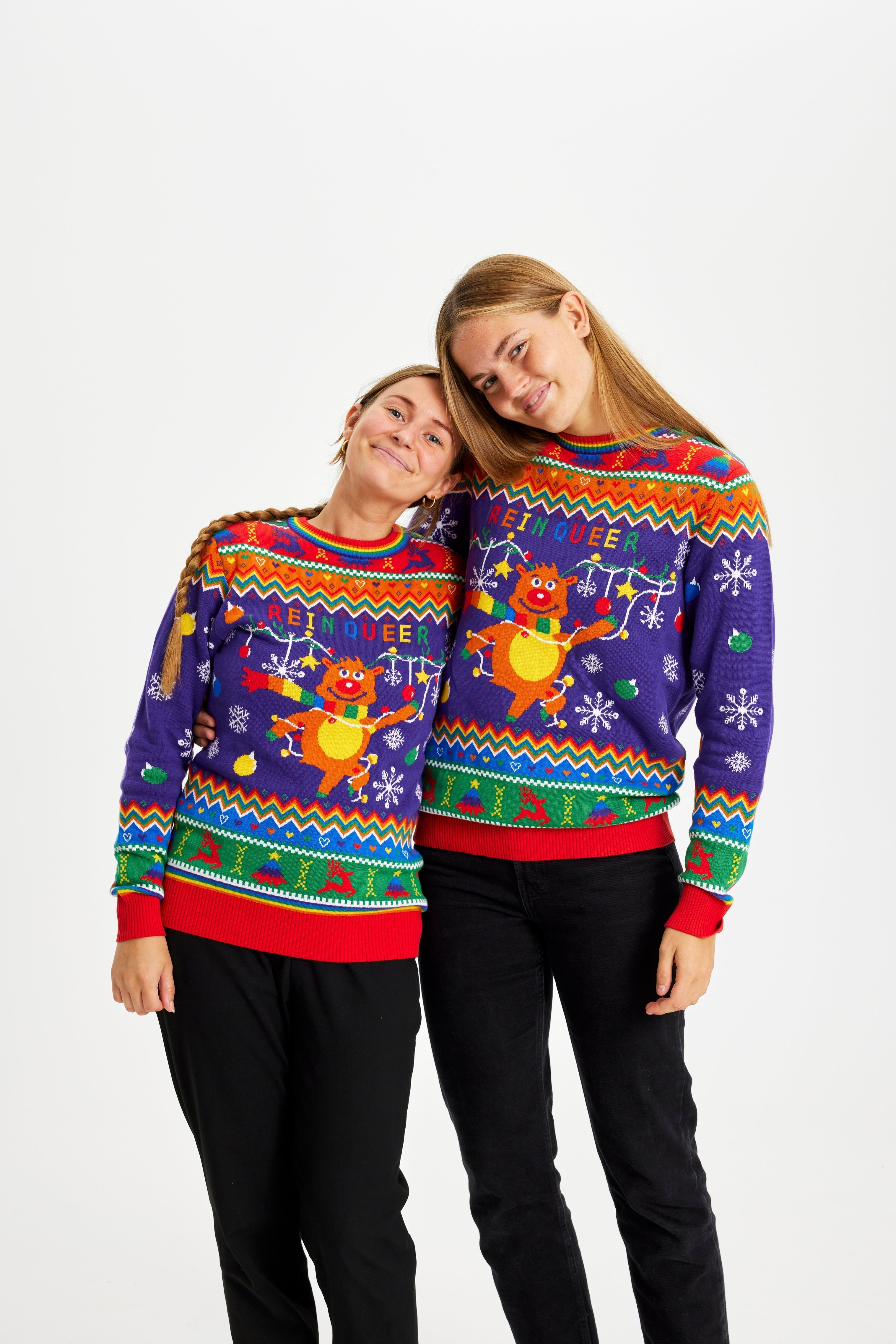 Deux femmes se tenant l'une l'autre, vêtues d'un pull de Noël aux couleurs de l'arc-en-ciel et portant l'inscription 