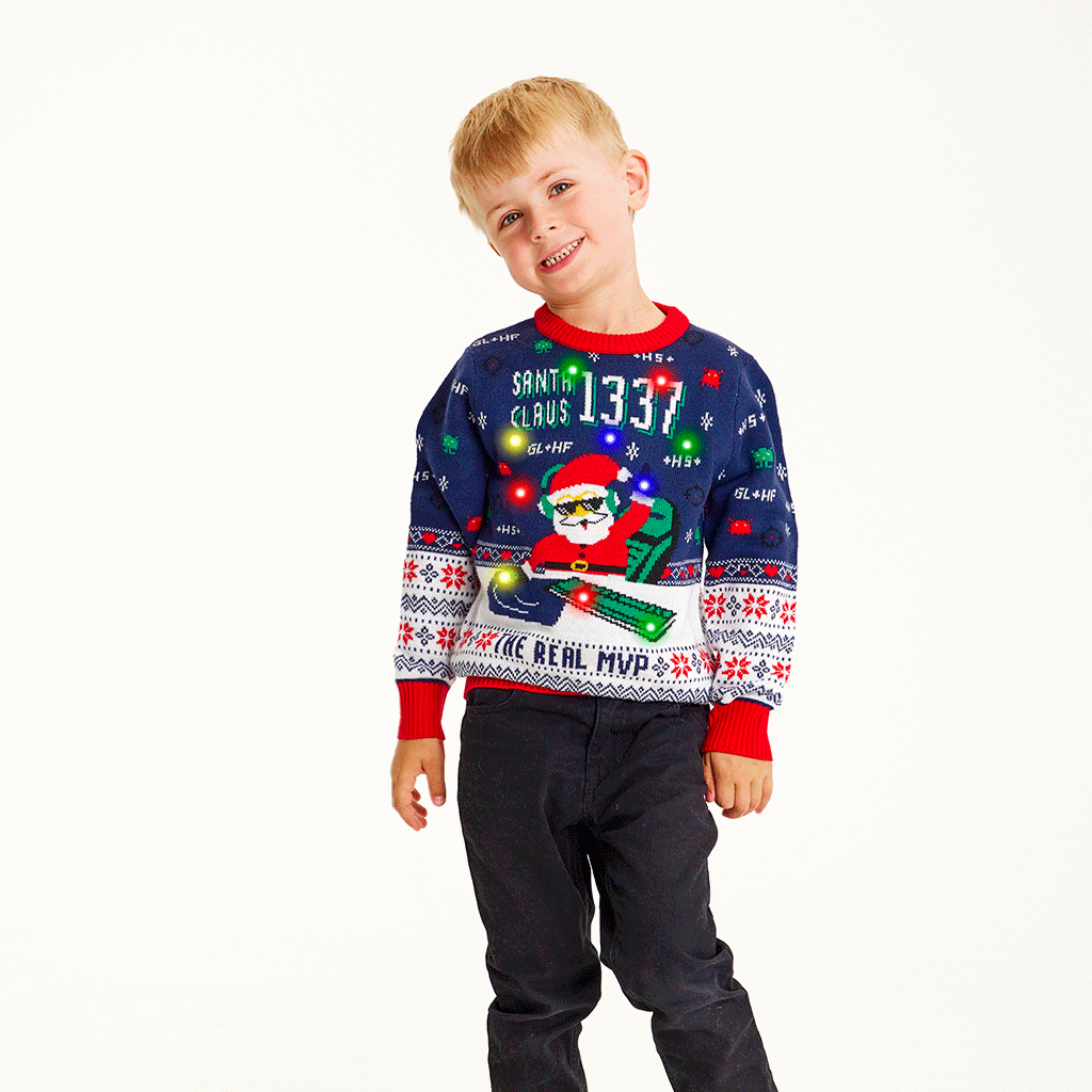 Un enfant souriant portant un pull de Noël bleu et rouge avec un Père Noël et des lumières.